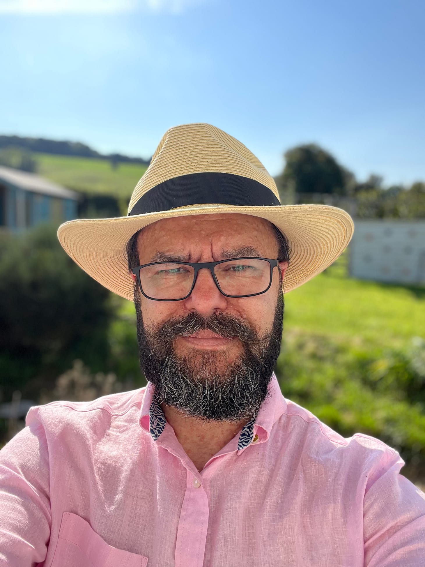 A bearded man with glasses grins coquettishly at the camera. He is wearing a pink shirt and a straw fedora.